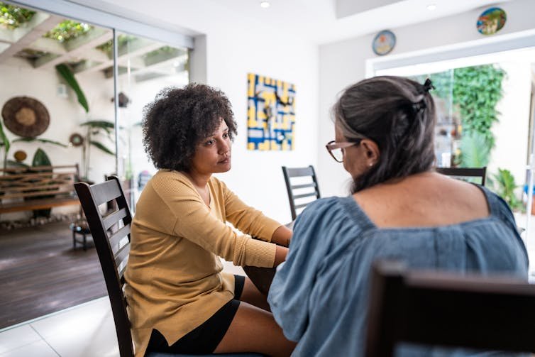 A young woman in a tan sweater and an older woman with gray hair sit at a table in a light-filled room.